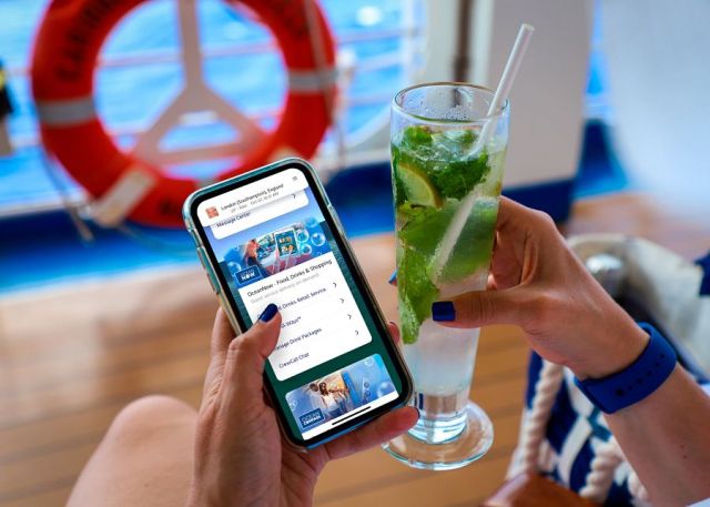 Hands with blue nail polish - left hand is holding a cell phone with Princess cruise app. Right hand is holding drink with mint and straw. Facing a orange life ring and sitting on a chair on wood deck.