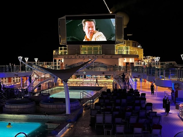 Pool deck of Sky Princess cruise ship at night. Lounge chairs are facing large screen over pools showing a movie. 