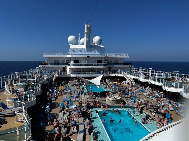 View looking over two main pools on Sky Princess cruise. People are on the deck and in the pools with blue skies.