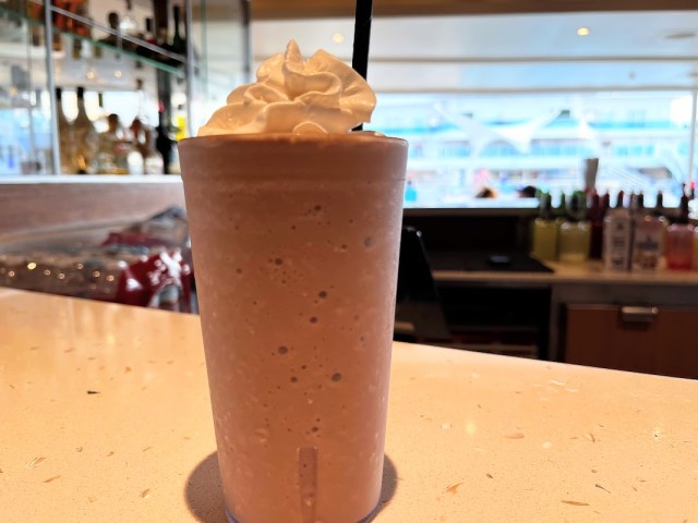 Chocolate frozen drink in plastic tumbler with whipped cream on top and black straw inserted. In faded out background is pool area of Sky Princess cruise ship.