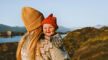 a mom who wanted to travel with baby standing in the mountains with her infant, both wearing beanie hats