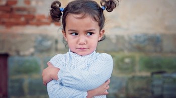 an angry toddler with pigtails crosses her arms in front of a brick wall