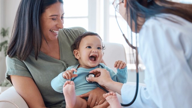 a baby smiles at a doctor using a stethoscope, sitting on mom's lap at the doctor's office