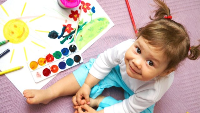 a little girls with pigtails sits by a painting with a sun and paints, toddler activities