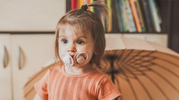 a toddler with two pacis in her mouth looking very resistant to pacifier weaning