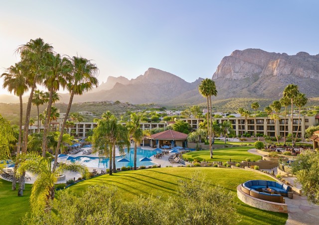 Resort view with green grass in the foreground, image of pool and blue waters in the middle and the mountains n the background against a pale blue sky.
