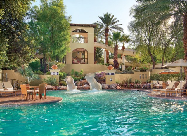 Pool area at a resorts showing daybeds, umbrella, and two water slides flowing into the pool. 