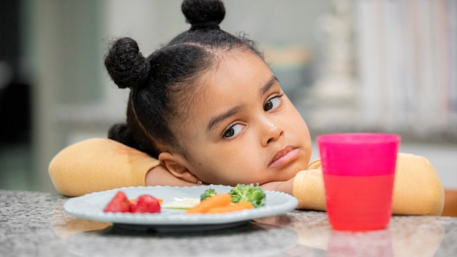 little girl resting her head on the table unhappily looking at a plate of food for a story on picky eater mistakes