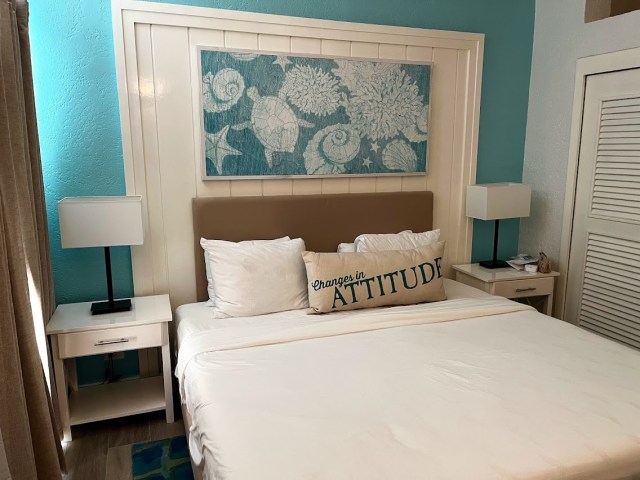 Looking at bed with Changes in Attitudes pillow on it. Above bed is art work of turles and walls are turquoise. Two night stands on either side of the bed with lamps on them. 