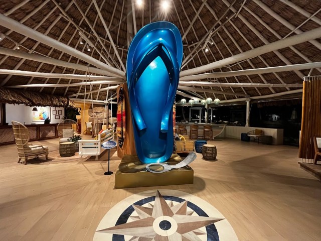 View of open air lobby with thatched roof, compass design on floor, large blue flip flop and sliver pop top sculpture