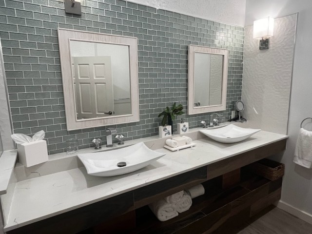 Bathroom vanity that extends from one wall to the other. Two mirrors hang over two sinks. Wall behind mirrors has glass subway tiles while vanity under sinks is dark brown and has white towels inside. 