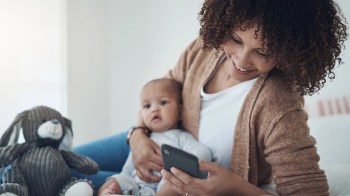 A mom on her bed holding her baby while looking on her phone at one of the best apps for moms
