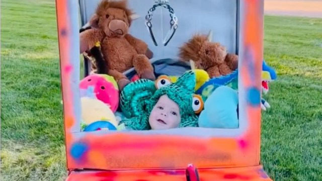 a baby in a Halloween stroller costume that makes them a stuffie inside a Claw arcade machine