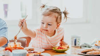 A toddler girl with pigtails reaching for more food on the table for a story on toddler dinner ideas