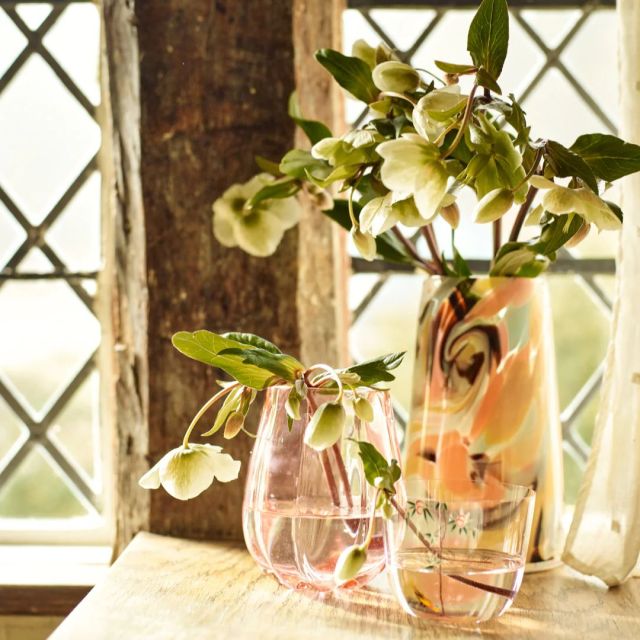 two multicolored glass vases filled with flowers on table in front of a sunny window