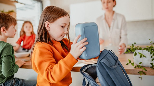 a little girl putting her school lunch in her backpack looking like she probably won't eat it